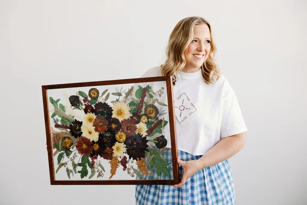 A woman poses with a large scale framed pressed flower bouquet preservation piece.