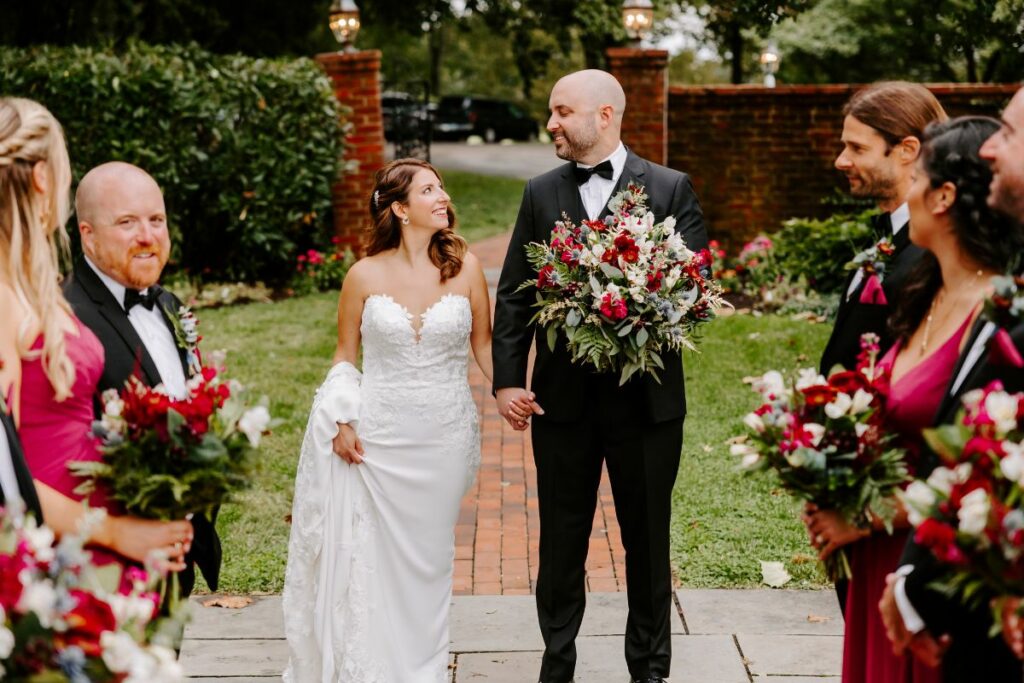 Bride and Groom look at each other in between their wedding party, all holding jewel tone wedding flowers at Brittland Estates.