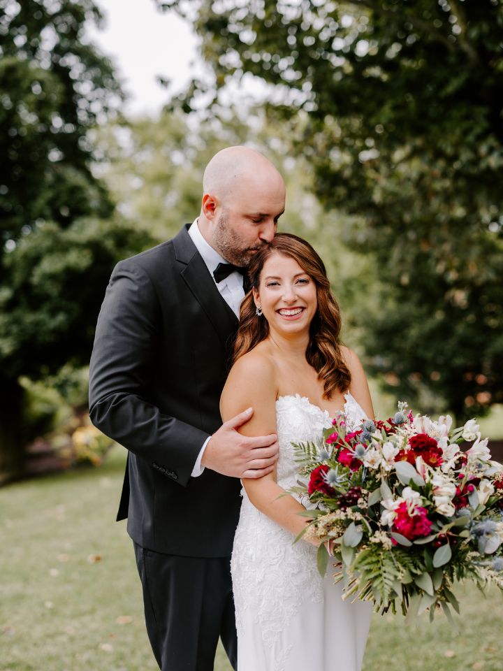 Groom kisses top of bride's head while she looks at the camera, holding her large bouquet of maroon and blue jewel tone flowers.