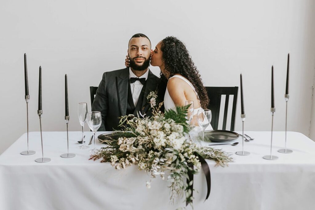 Bride kisses groom on the cheek while seated at the sweetheart table, her green and white bouquet on the table with taper candles.