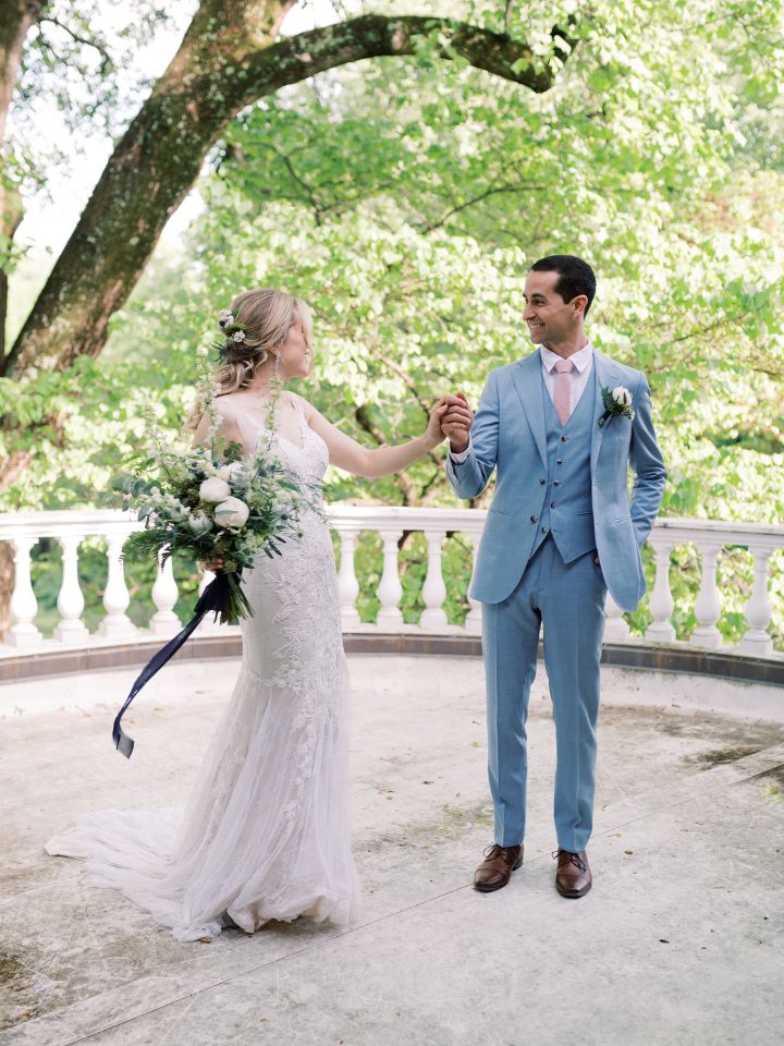 Bride and Groom twirl on a patio under the trees holding blue and white wedding flowers.