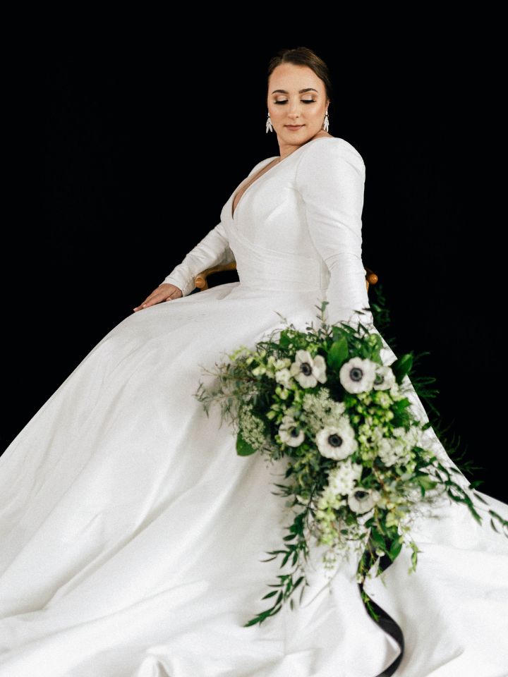 Black and White Florals Bride in white dress sits in front of black wall, holding a white and green wedding bouquet down to her side with trailing black ribbon.