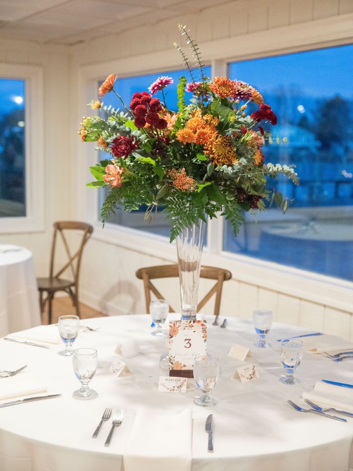 Guest table at a wedding reception has a tall glass pedestal with autumn wedding flowers in it.