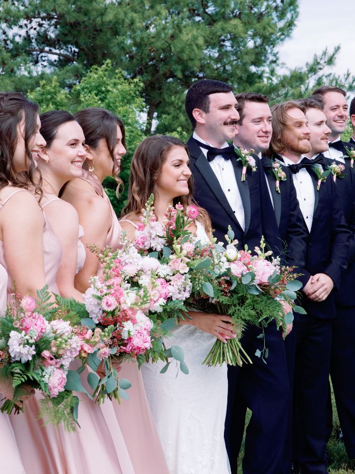 Groom and groomsmen in black tuxes line up next to the bride and bridesmaids.