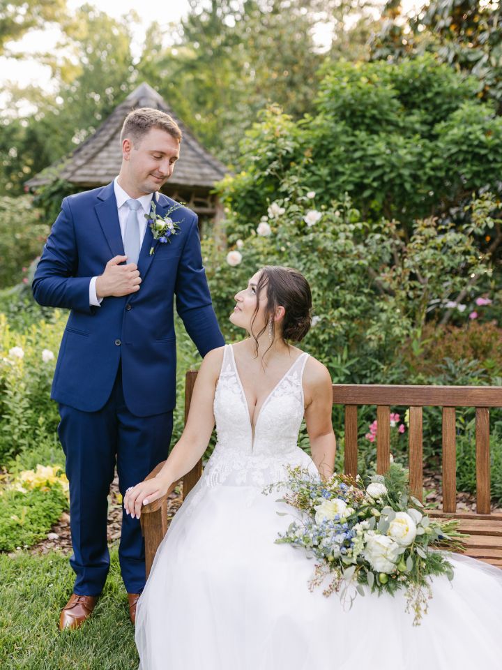 Bride sits on a garden bench holding her bouquet, looking up at her groom.