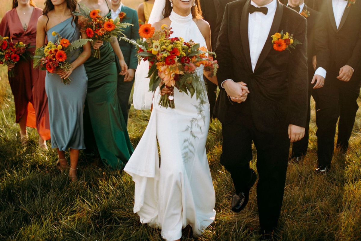 Thalia colton close up Bride and groom walk with bridal party behind them holding orange flower bouquets and wearing orange flower boutonnieres