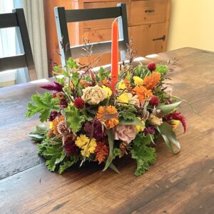 A table centerpiece flower arrangement in fall colors with a taper candle sits on a wooden dining room table.