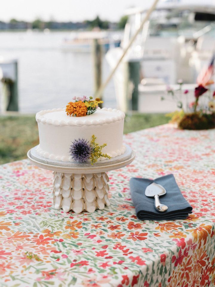 A small wedding cake sits on a textured stand on top of an orange and pink floral tablecloth, with edible florals decorating the icing.