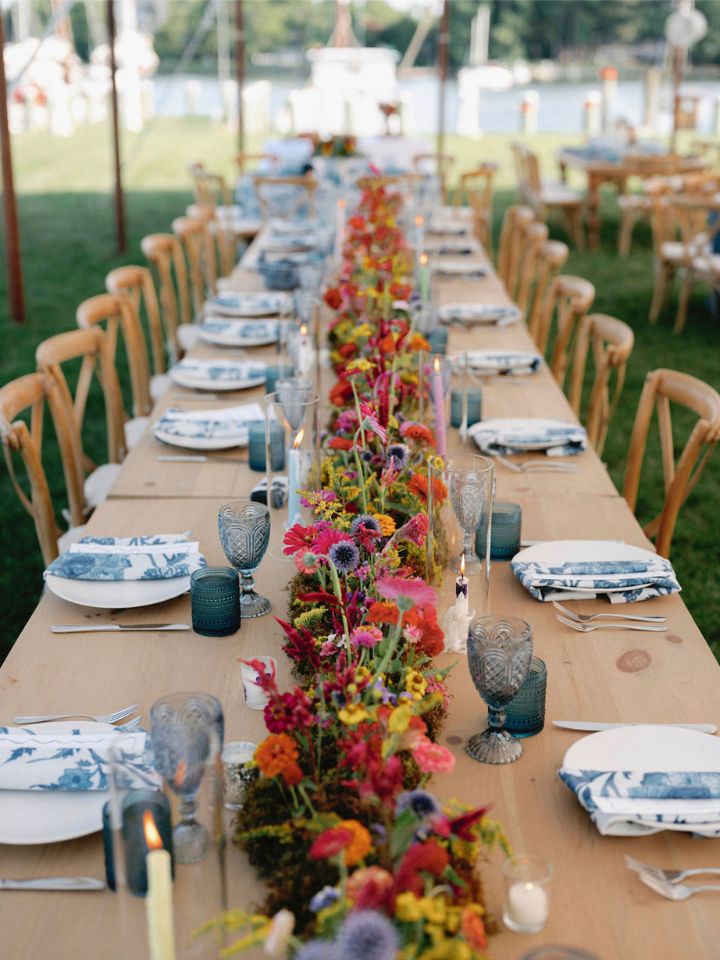 Looking down a wedding party table at an outdoor wedding reception, made up of three tables placed end to end.  There is a long floral arrangement with moss and bright flowers down the entire center.