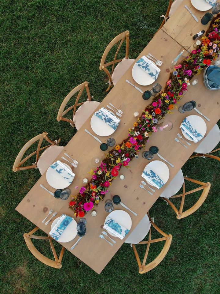 Overhead view of a light wood harvest table on grass with crossback chairs, place settings, and a wildflower moss meadow runner centerpiece.