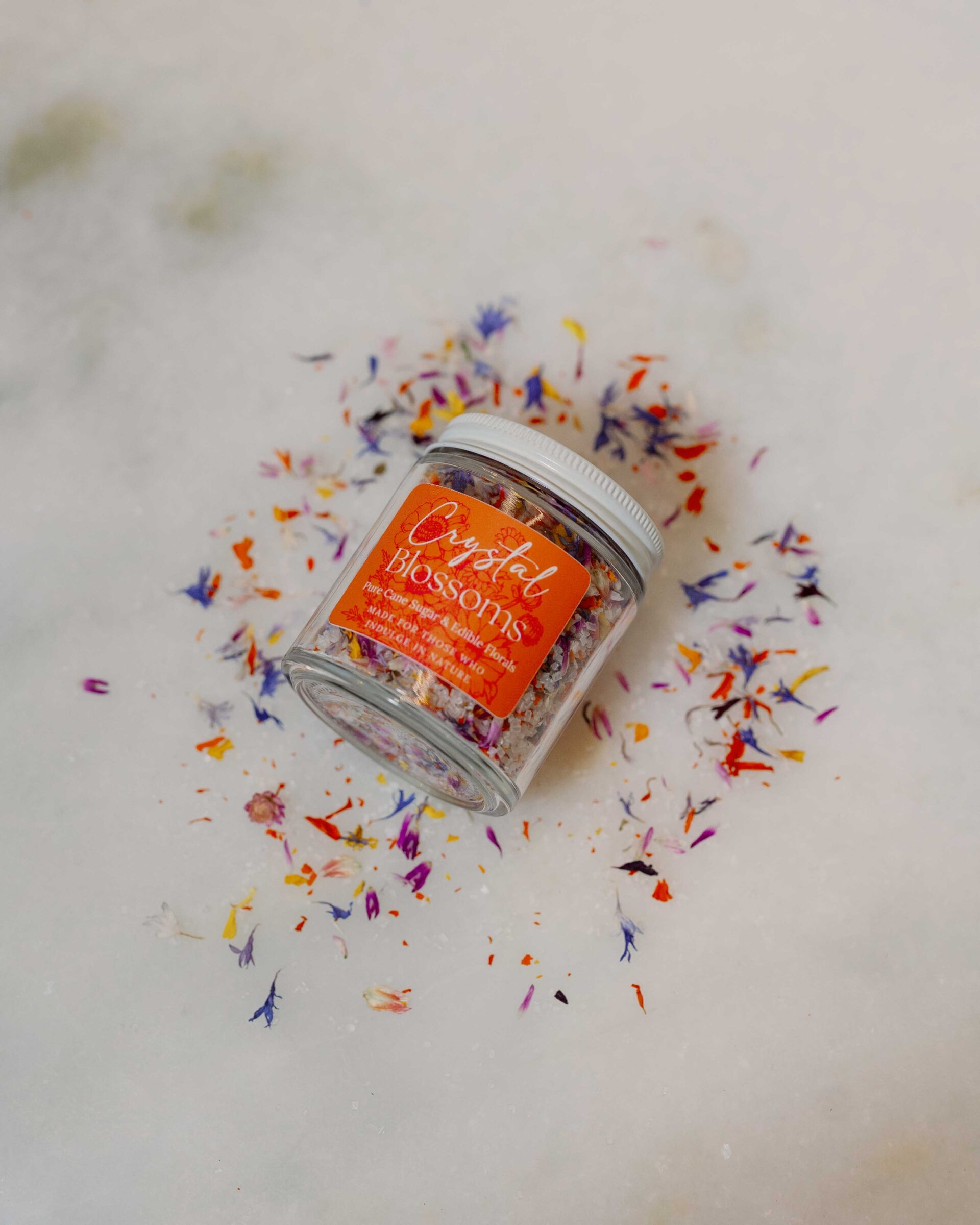 Jar of edible floral sugar on a white countertop surrounded by petals and sugar.