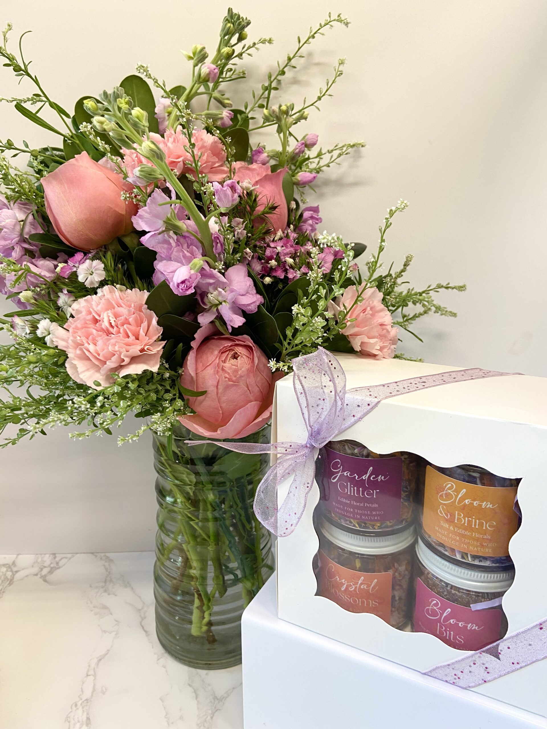 A flower vase stands behind a gift set of four jars of edible florals.