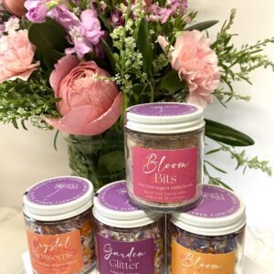 Jars of edible floral sugars, petals, and salts sit in front of a pink and purple floral arrangement.