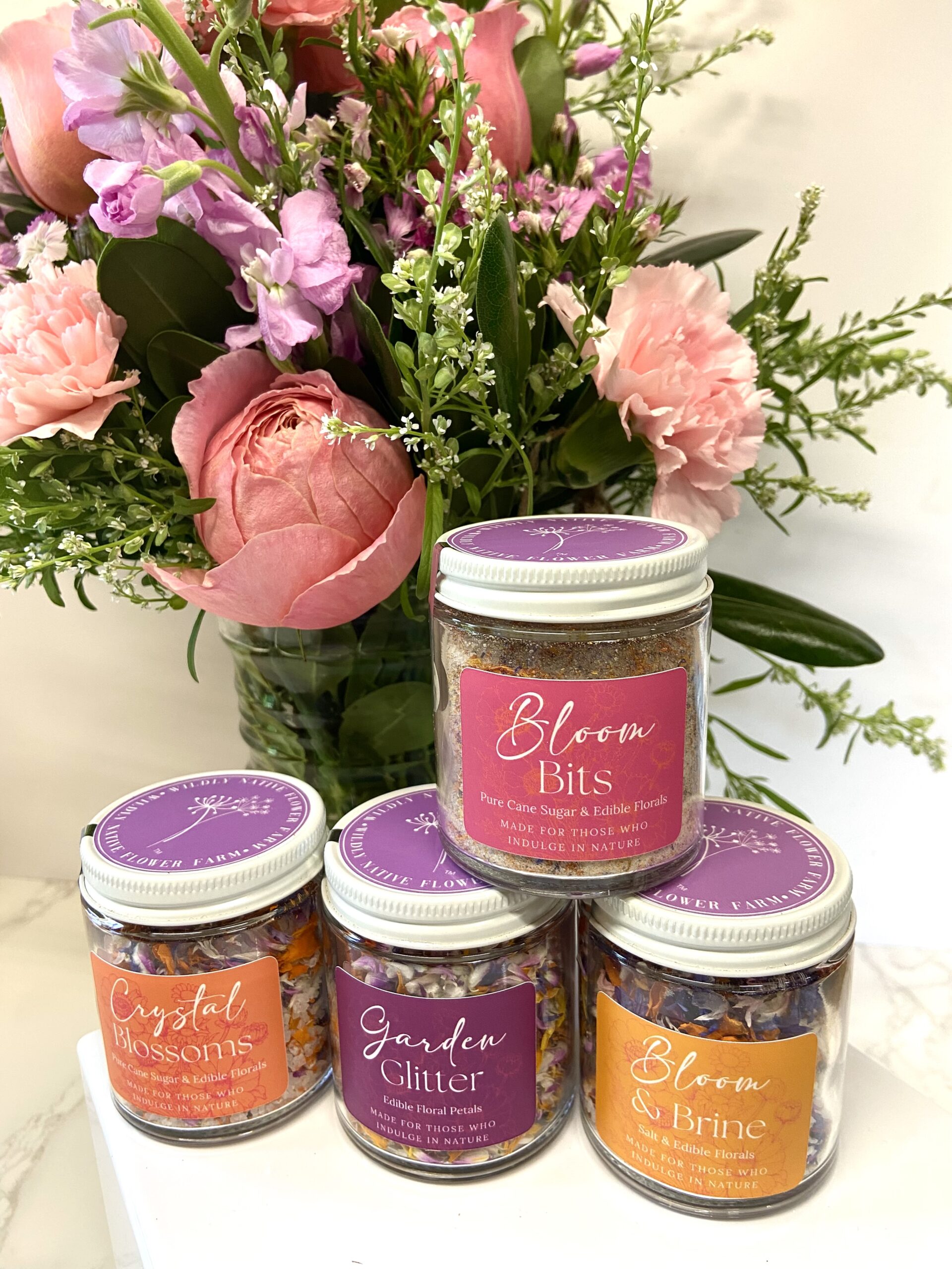 Jars of edible floral sugars, petals, and salts sit in front of a pink and purple floral arrangement.