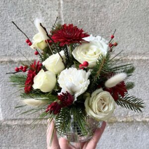 Photo shows a hand holding a small disco flower vase arrangement up in front of a cinder block wall, full of Christmas color flowers and evergreens.