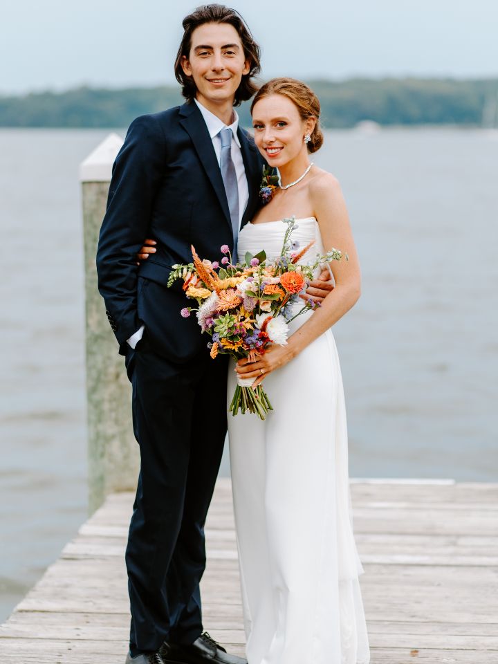 Bride and groom pose on a dock, holding her flower bouquet towards camera.