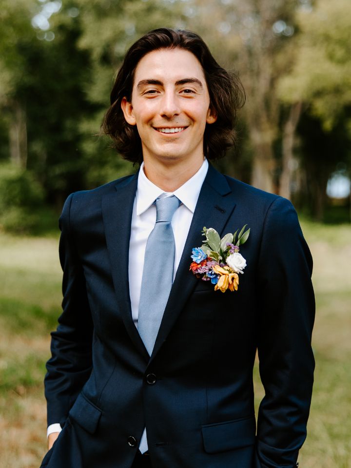 Groom in black suit and light blue tie smiles while wearing a flower pocket square.