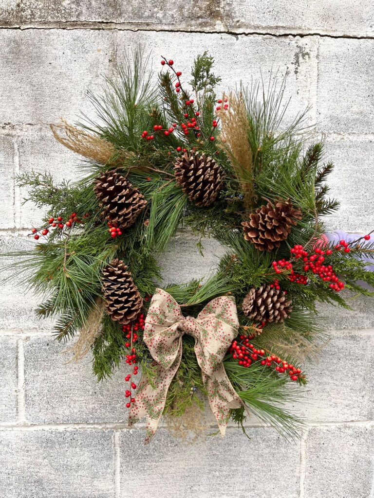 A finished Christmas wreath is held in front of a cinder block wall, and has evergreens, pine cones, red berries, grasses, and a bow.