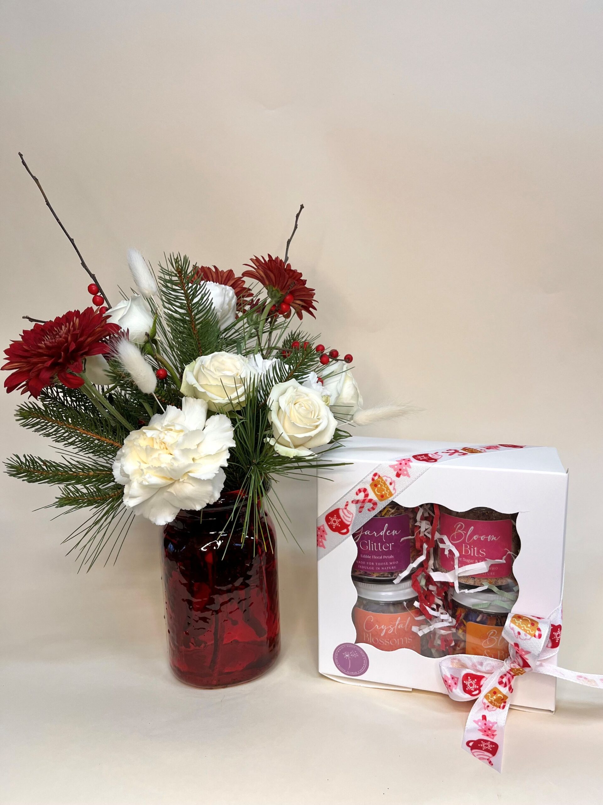 A red vase with Christmas flowers sits next to a white boxed gift set of edible floral petal sugars and salts.