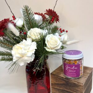 A vase or Christmas flowers sits next to a wood block with a jar of edible flower petals sitting on top.