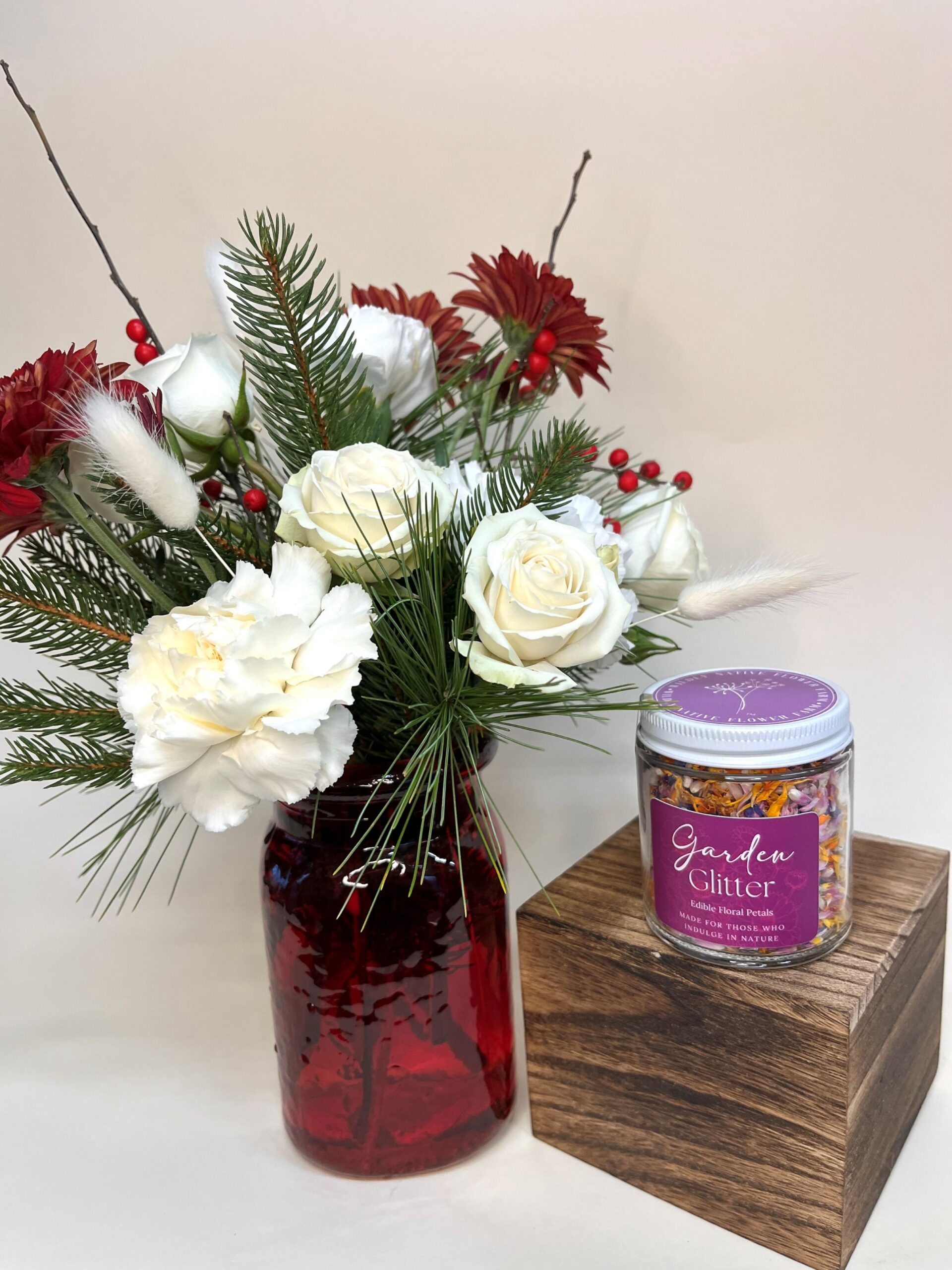 A vase or Christmas flowers sits next to a wood block with a jar of edible flower petals sitting on top.