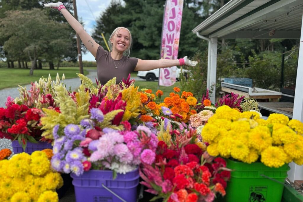 Lizzy stands with arms open behind a gator full of cut flowers from the flower farm