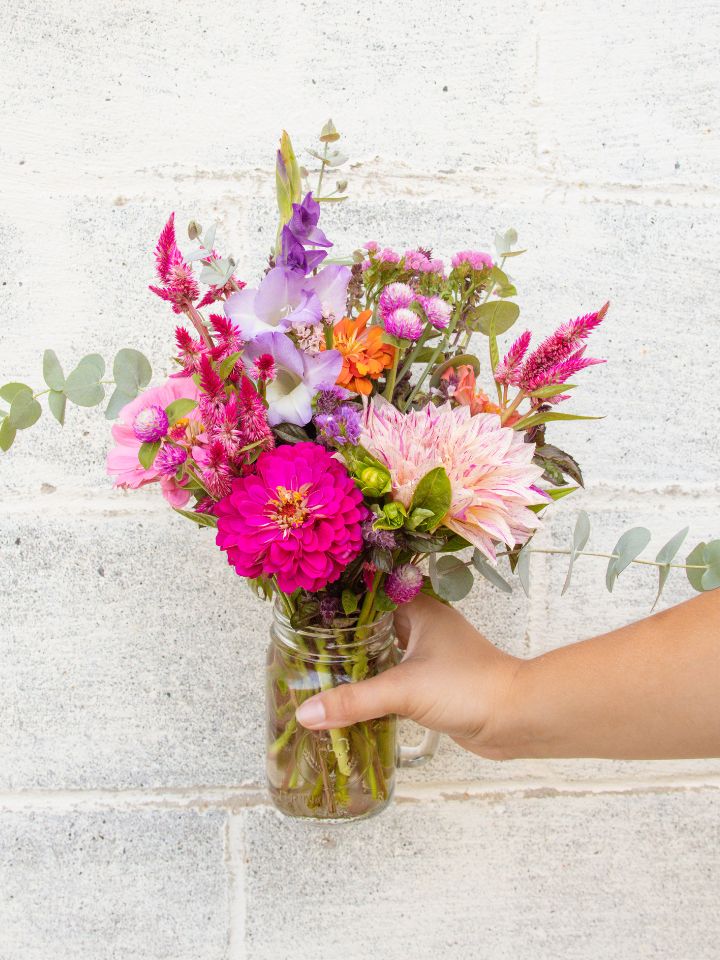 Hand holds a vase of cut flowers in front of a cinder block wall.