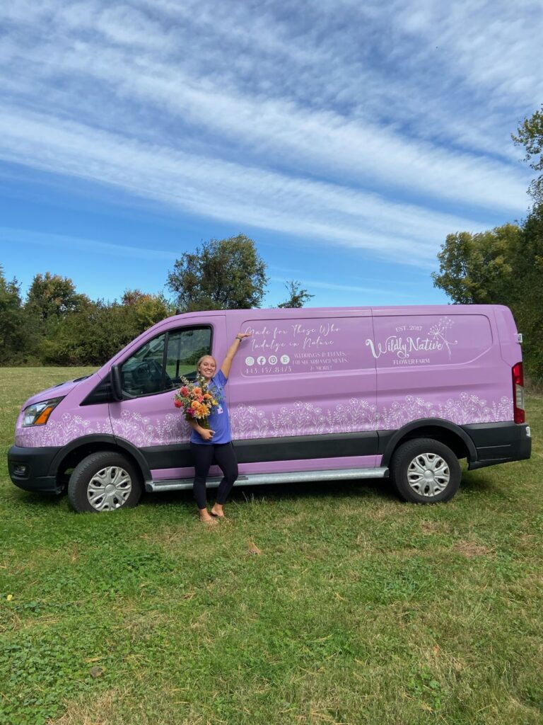 Lizzy holds a flower bouquet in front of our purple delivery van.