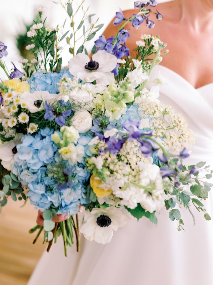 Closeup of bridal bouquet with white anemones, blue hydrangea, and pops of yellow ranunculus.
