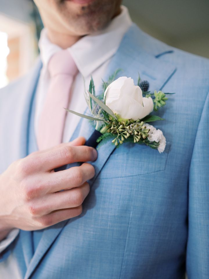 Groom holds white floral boutonniere on his pale blue suit with coral pink tie.
