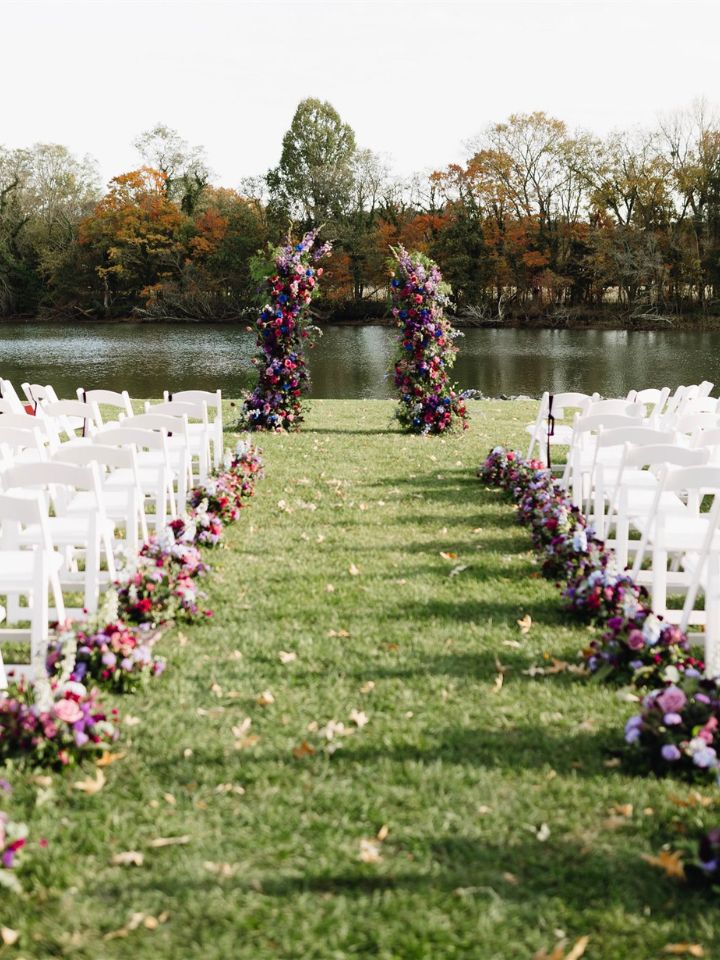 Looking down a grassy aisle at a waterfront wedding ceremony at the Oaks, with purple and blue flower arrangements down the aisle and at the end.
