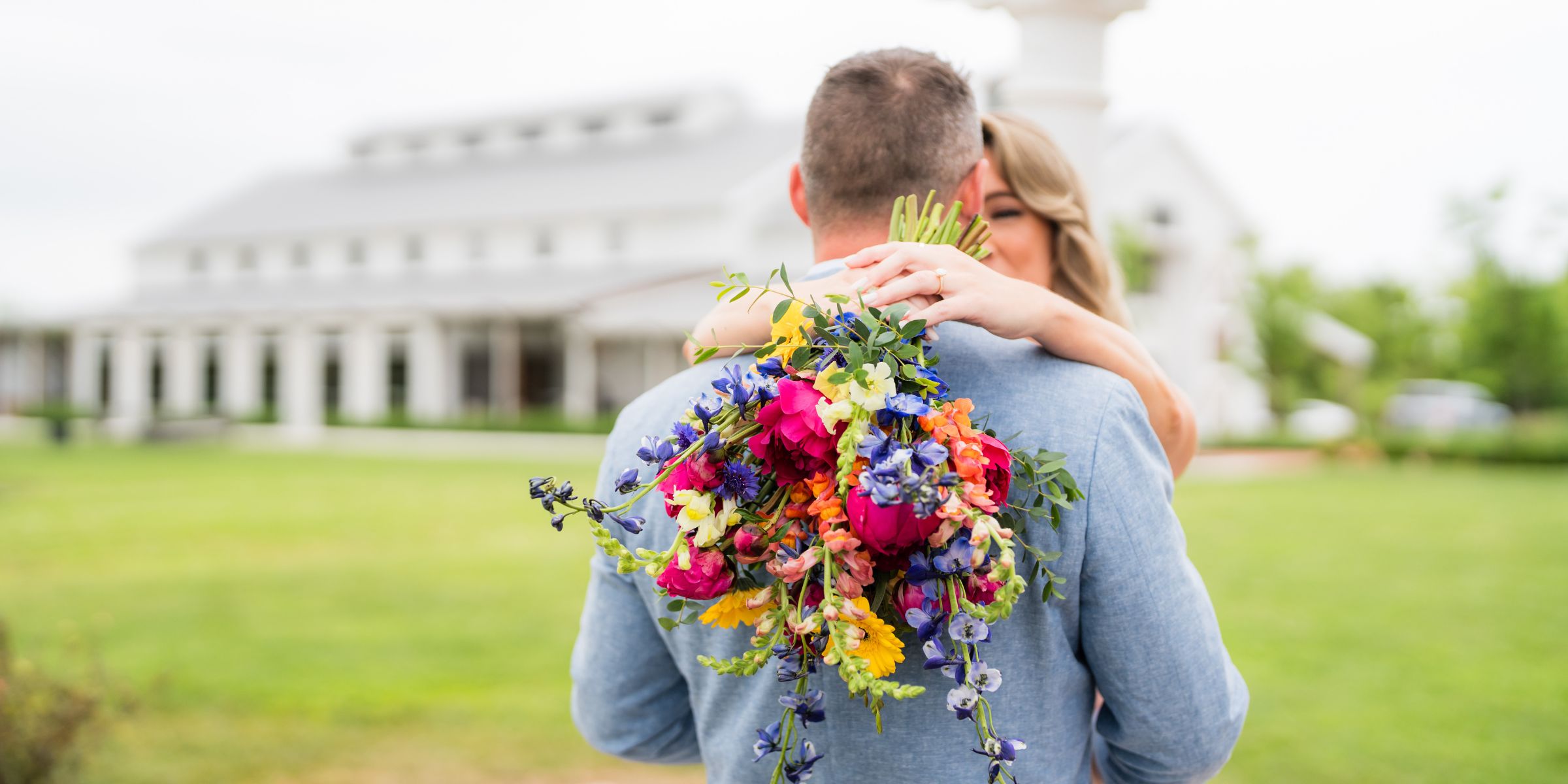 Groom hugs bride, she has her bouquet behind him showing to the camera, Kent Island Resort Farmstead in the background.