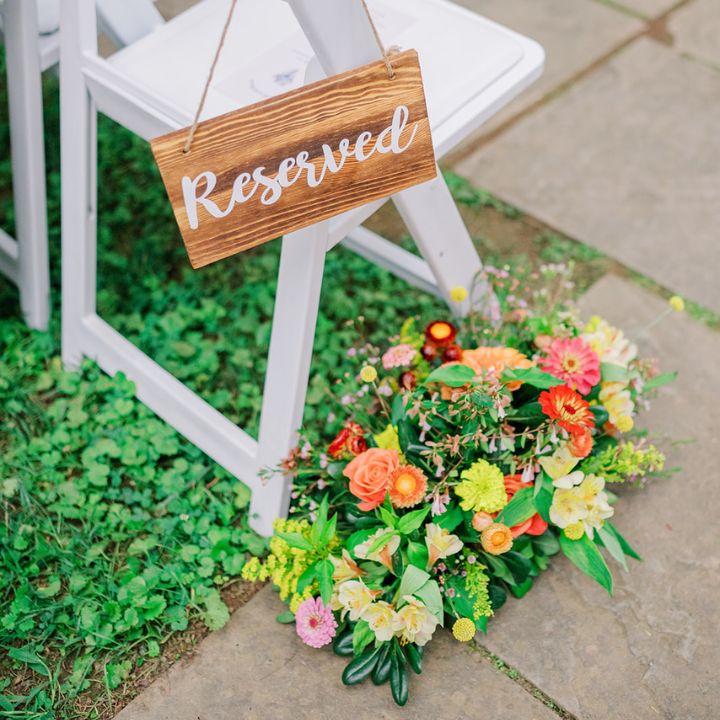 Orange, yellow, and pink flower arrangement at base of chair down the aisle at a wedding ceremony with a reserved sign.