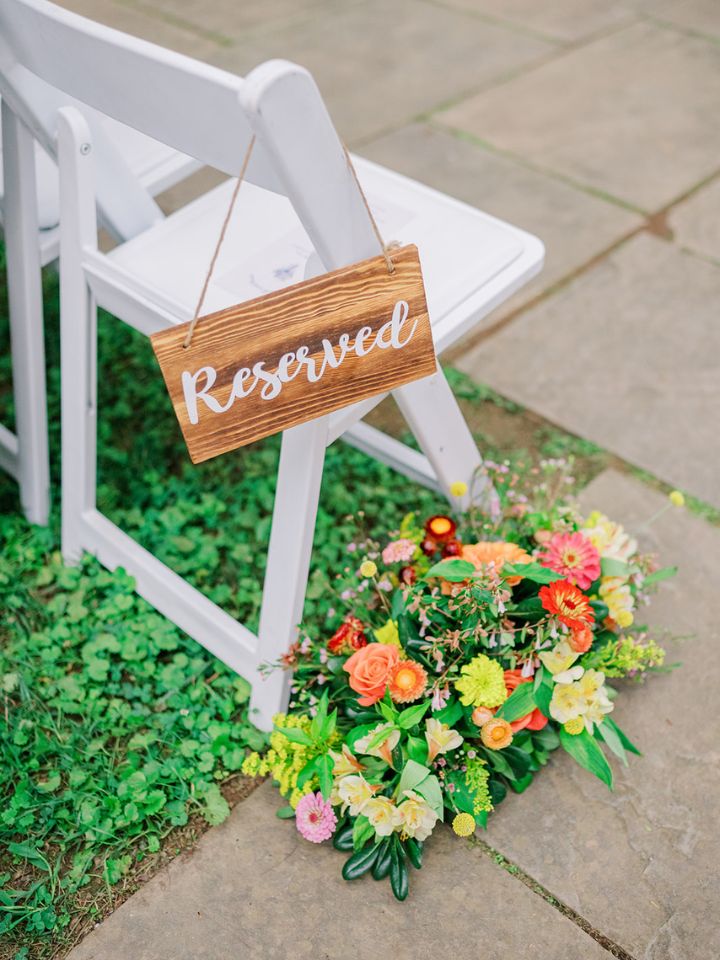 aisle florals green yellow orange A white chair sits in the grass with a reserved sign and floral arrangement at base.