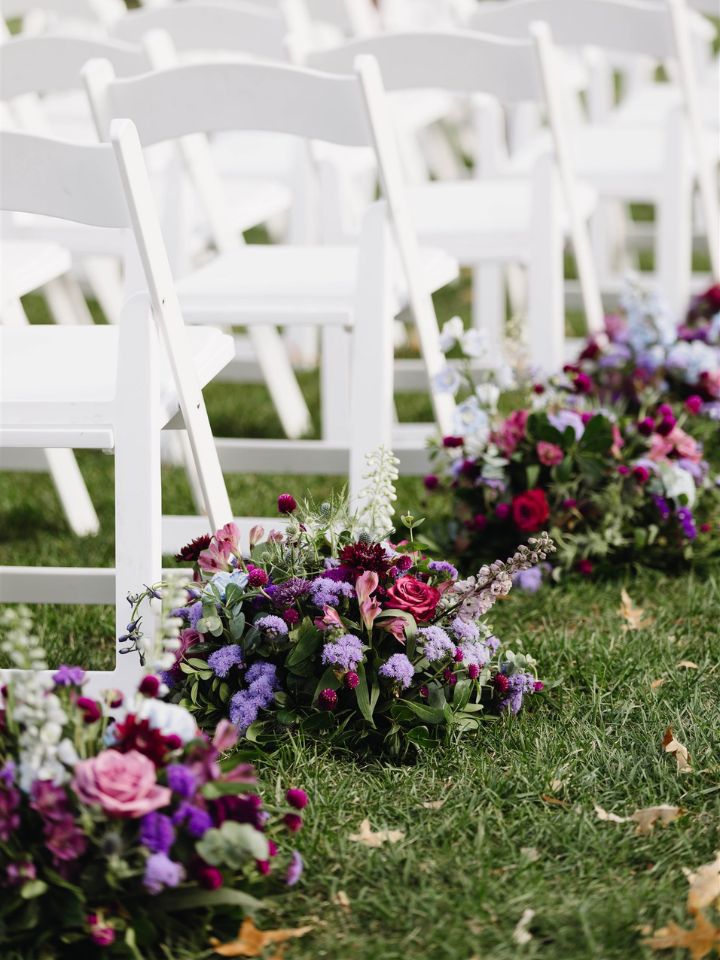 aisle flowers purple purple and blue flower arrangements line the aisle of white chairs at a wedding ceremony.