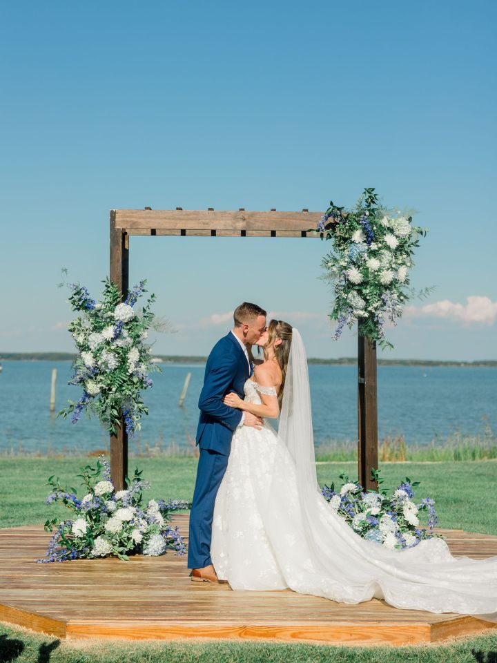 Bride and groom kiss under a wooden arch at the waterfront wedding ceremony at the Wylder Hotel, with blue and white florals.