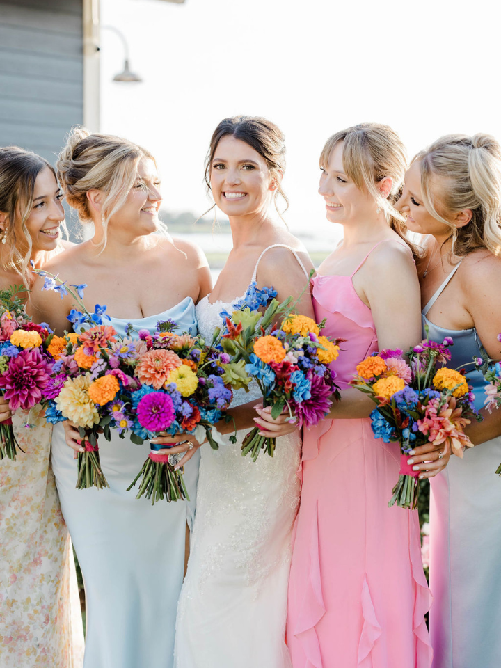 bouquet page main Bride stands with bridesmaids in pastel colorful dresses, all holding bouquets in bright summer flowers.