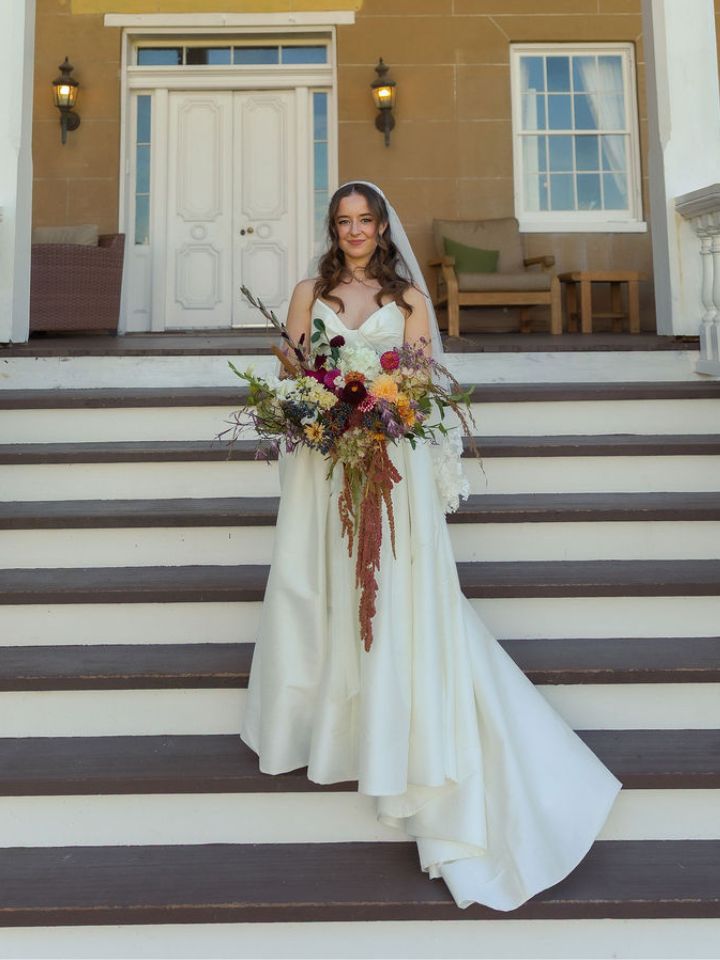 cascading classic jillian Bride stands on steps of Piney Grove holding a rich jewel tone fall bridal bouquet with cascading amaranth.