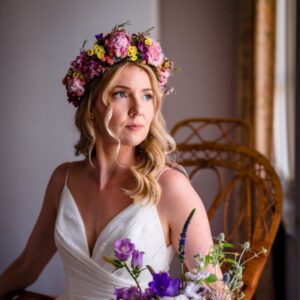 cassidy crown sq Bride wears a pink spring flower crown while posing seated in her wedding dress.