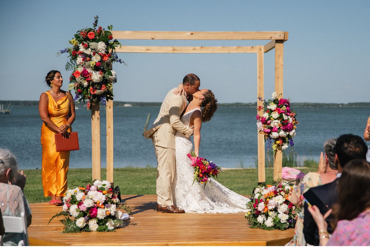 Couple kisses under pale wood chuppah with flowers.