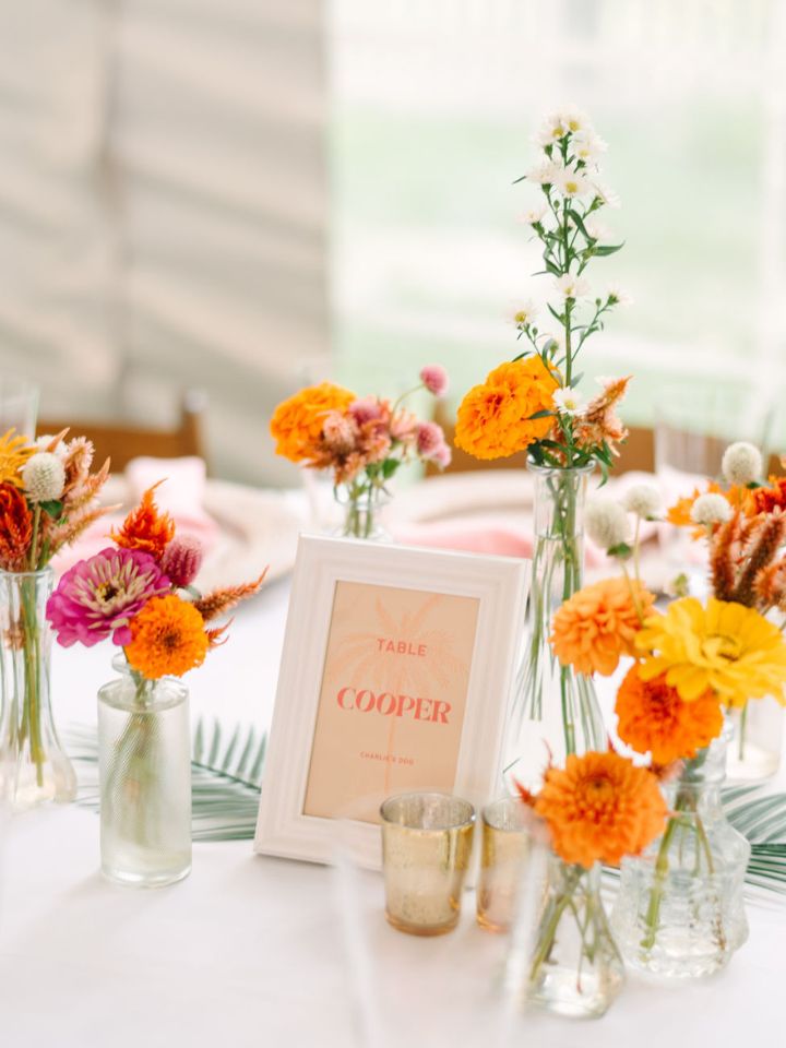 Colorful orange, pink, and yellow flowers in glass bud vases sit on a white tablecloth.