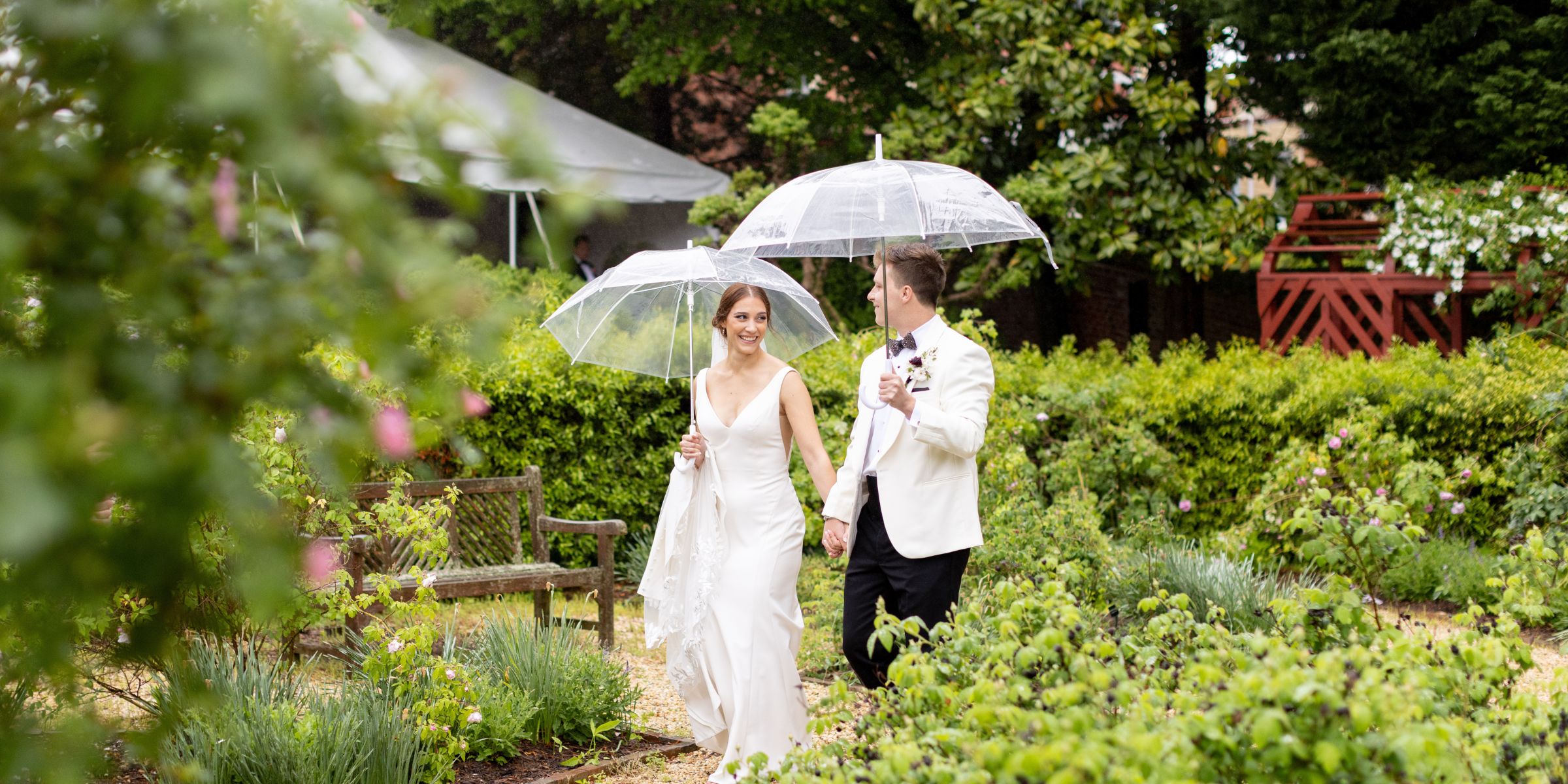 Bride and groom use large clear umbrellas while walking in the gardens at the William Paca House.
