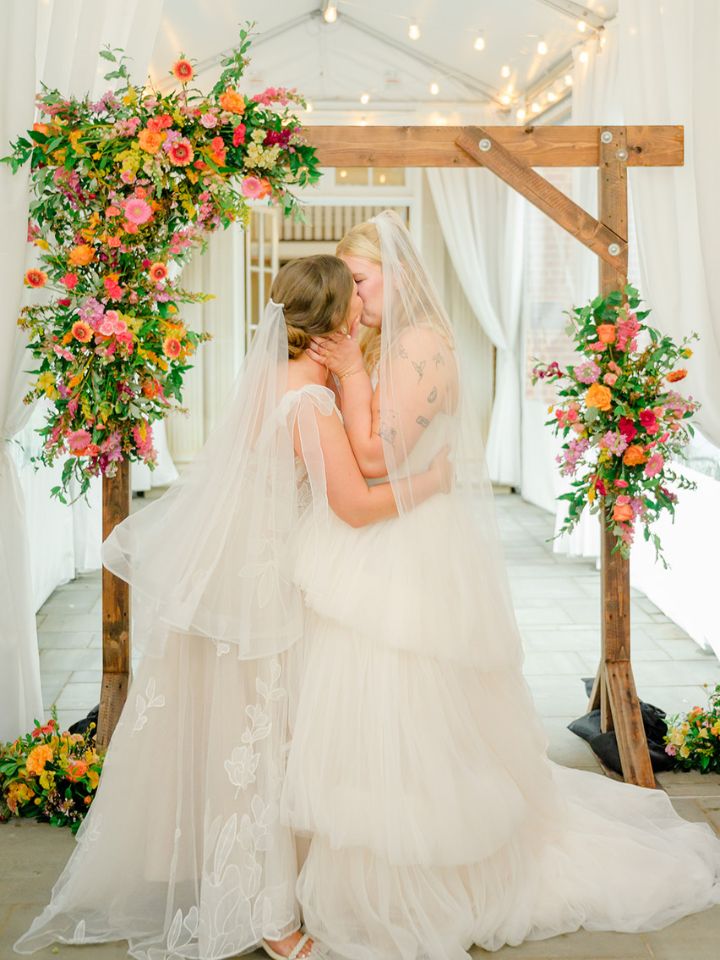 Brides kiss under wedding ceremony arch with colorful summer florals.