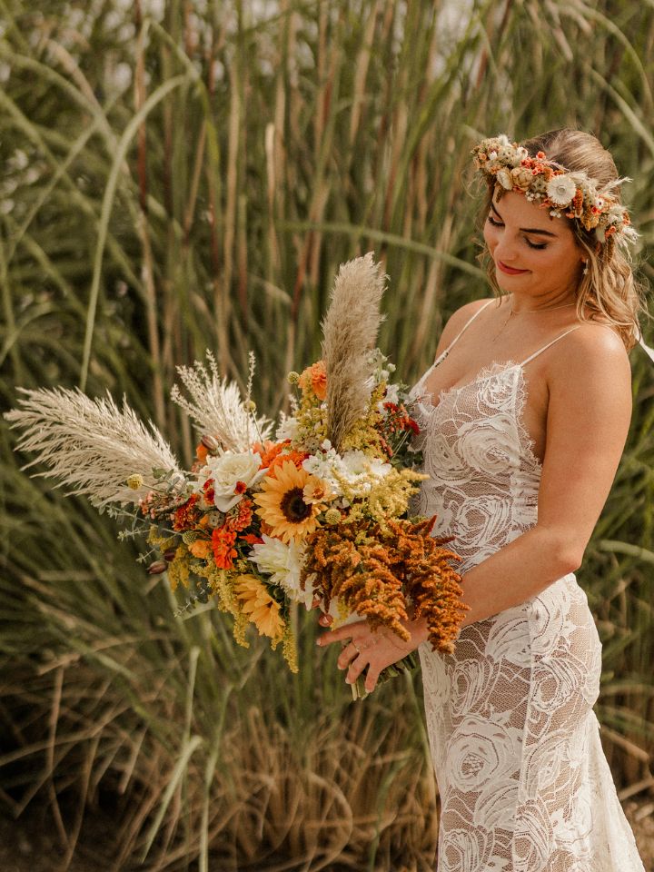 danielle showstopper organic Bride stands in front of waterfront grasses holding her bridal bouquet with sunflowers and grasses.