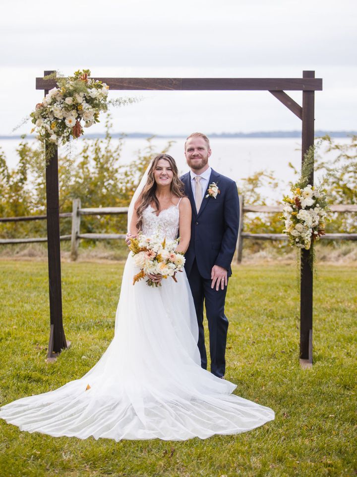 Bride and groom smile at camera, posing next to arch with flowers.