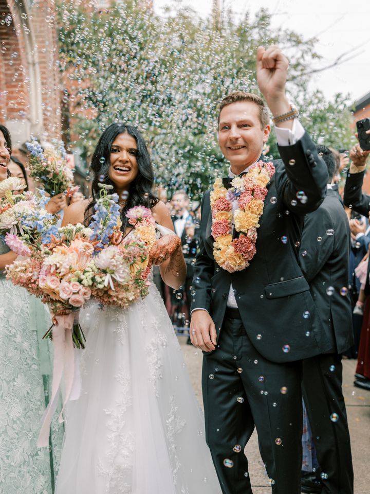 jai mala flower necklace Bride and groom celebrate on the sidewalk after wedding ceremony, with bubbles and peachy floral jai mala necklaces.