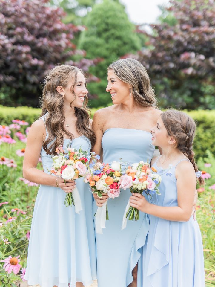 mini bridesmaids Three women hold miniature floral bouquets in bright colors, wearing pale blue dresses at a wedding.