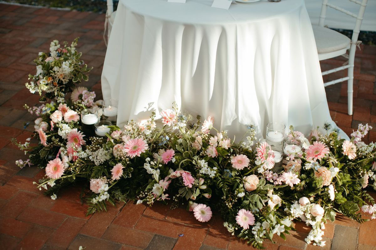 White linen table on brick patio with white and pink florals at base.