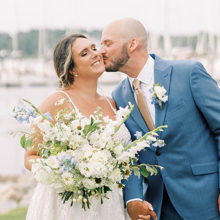 Groom in blue suit kisses bride holding her white and blue bouquet