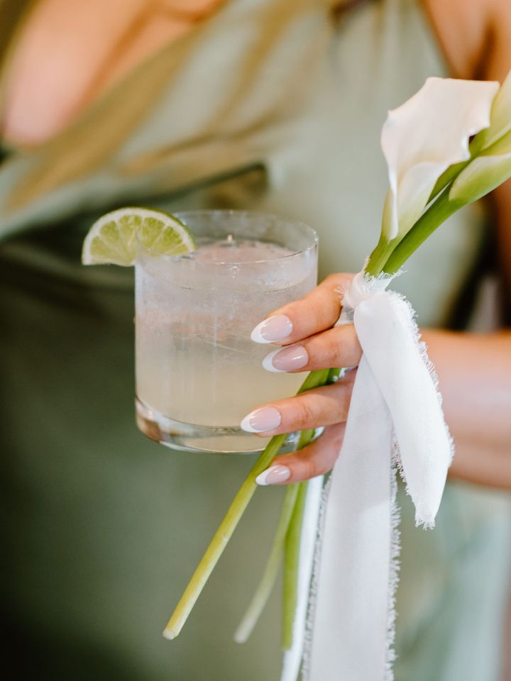 Hand holding a cocktail with lime slice and her mini calla lily bouquet.